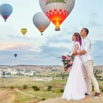 a man and woman standing on a hill with hot air balloons in the background