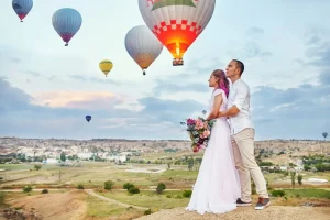 a man and woman standing on a hill with hot air balloons in the background