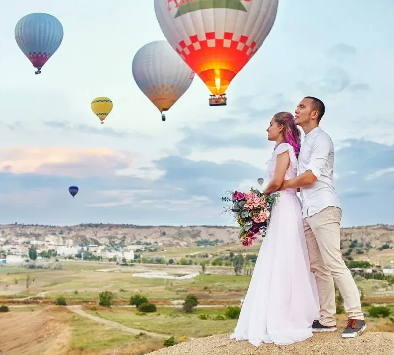 a man and woman standing on a hill with hot air balloons in the background