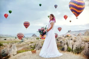 a woman in a white dress holding a bouquet of flowers in front of a group of hot air balloons